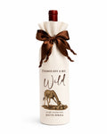A wine bottle wrapped in a premium calico fabric bottle bag with a dark brown satin bow at the neck, featuring a giraffe illustration and the text 'THINGS GOT A BIT Wild SOUTH AFRICA'.